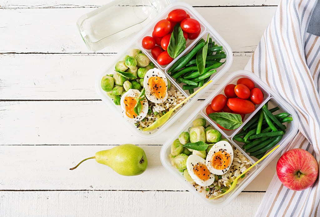 Meal prep containers filled with boiled eggs, Brussels sprouts, green beans, cherry tomatoes, spinach, and rice on a white wooden table with a pear, apple, and water bottle nearby.