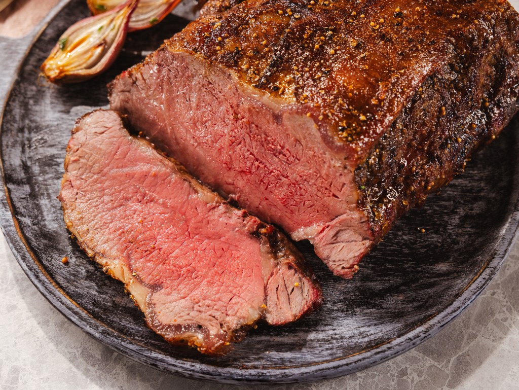 A perfectly cooked Certified Angus Beef® strip roast, sliced to reveal its tender, juicy interior, served on a rustic black plate with roasted shallots.