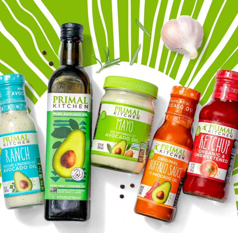 Assorted Primal Kitchen products featuring avocado oil, including ranch dressing, pure avocado oil, mayonnaise, buffalo sauce, and organic ketchup, displayed against a green background with garlic and herbs.