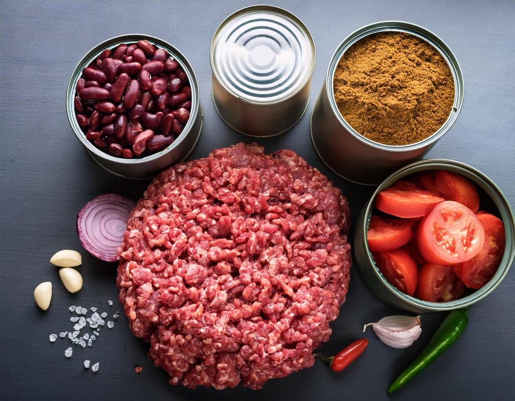 Fresh ground beef surrounded by ingredients for homemade chili, including canned red kidney beans, canned diced tomatoes, chili powder, garlic, red onion, sea salt, and chili peppers, arranged on a dark surface.