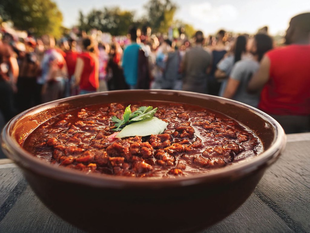 A large bowl of thick, hearty chili garnished with cilantro and sliced jalapeño, set against a blurred background of people at an outdoor festival.
