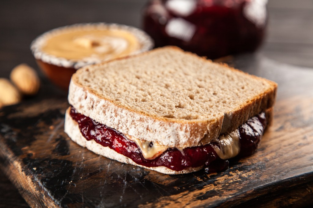 Close-up of a peanut butter and jelly sandwich made with whole wheat bread, placed on a rustic wooden cutting board with blurred ingredients in the background