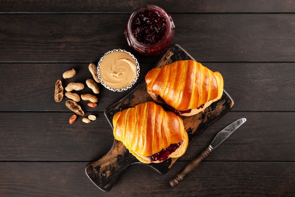 Freshly baked croissant sandwiches filled with peanut butter and jelly, served on a rustic wooden cutting board with jars of spread and peanuts
