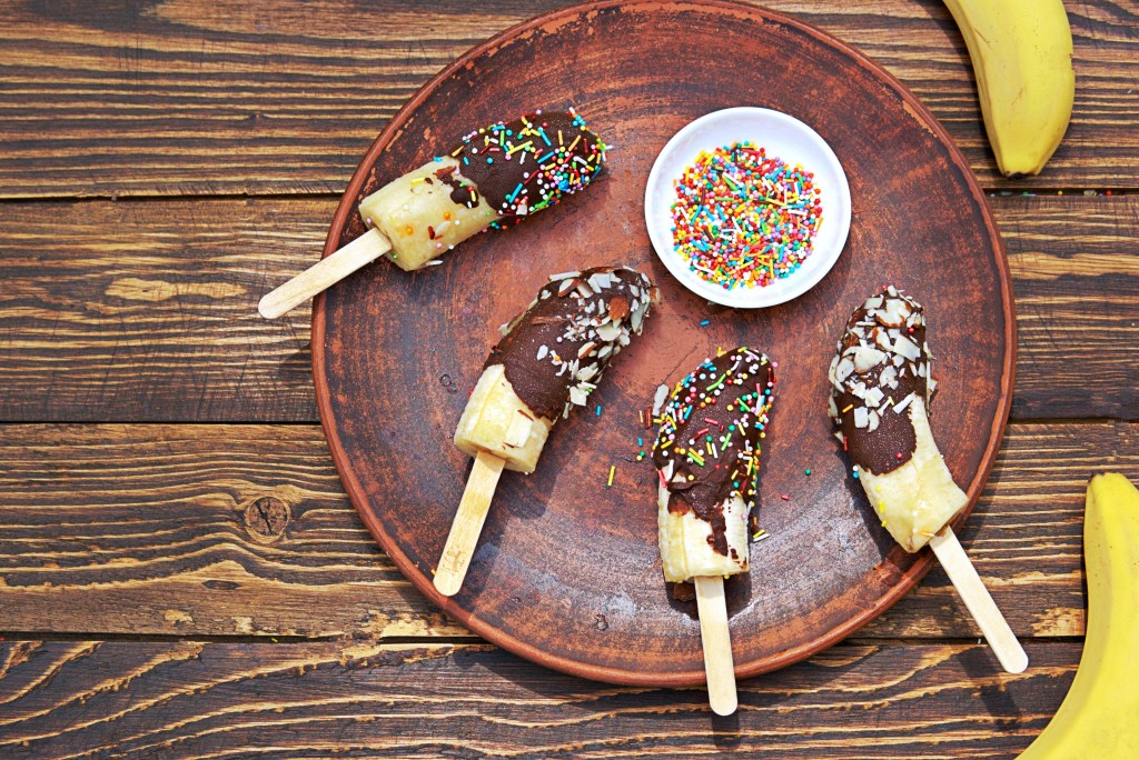 Frozen banana pops dipped in chocolate and topped with sprinkles and nuts on a brown ceramic plate with wooden background