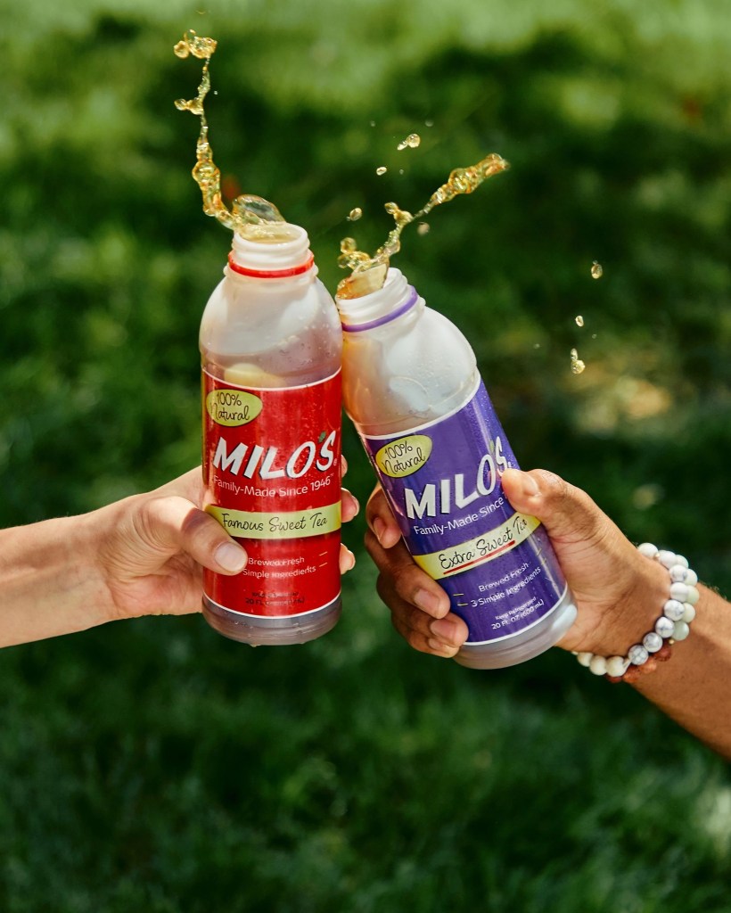 Two hands clinking Milo’s Famous Sweet Tea and Extra Sweet Tea bottles with tea splashing mid-air in a green outdoor setting