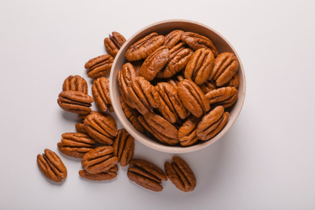 Top view of whole shelled pecans in a wooden bowl with scattered nuts on a white surface