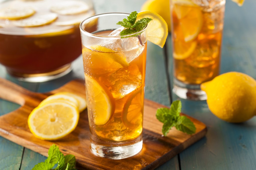 Tall glasses of iced tea with lemon slices and mint leaves on a wooden board with a pitcher of tea and fresh lemons in the background