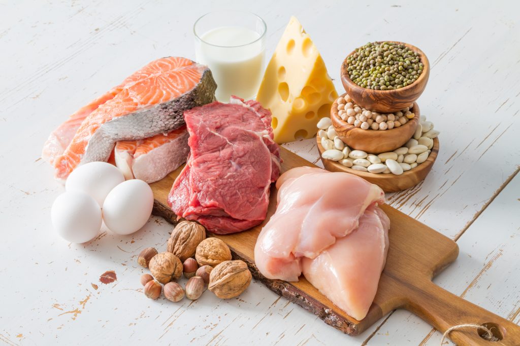 Assorted protein sources including raw salmon, beef, chicken breast, eggs, cheese, milk, beans, lentils, and nuts on a wooden board