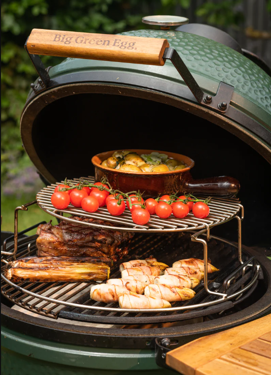 Big Green Egg grill with cherry tomatoes, bacon-wrapped chicken, roasted vegetables, and ribs cooking outdoors