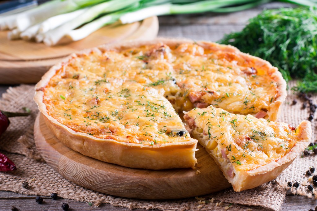Homemade quiche with a golden cheese topping and herbs, served on a wooden board with fresh dill and green onions