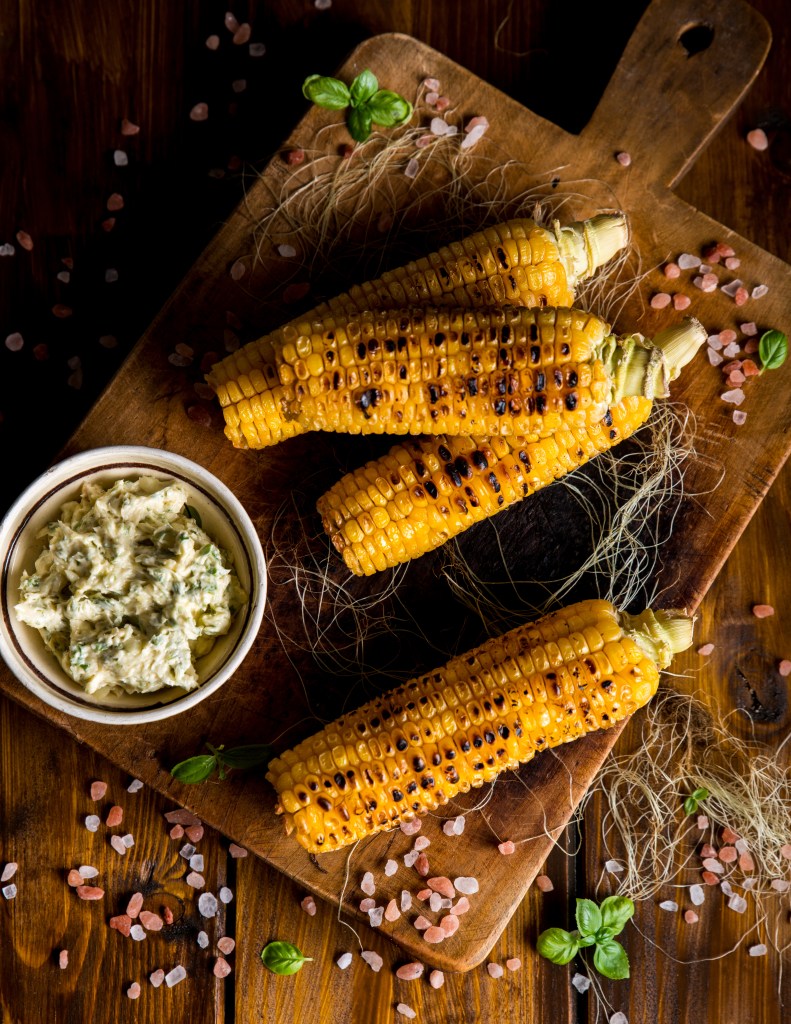 Char-grilled corn on the cob served on a rustic wooden board with a side of herb-infused butter and pink Himalayan salt