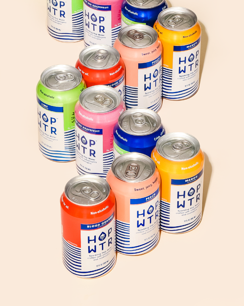 Assorted HOP WTR non-alcoholic sparkling hop water cans in multiple vibrant flavors neatly arranged on a light beige background
