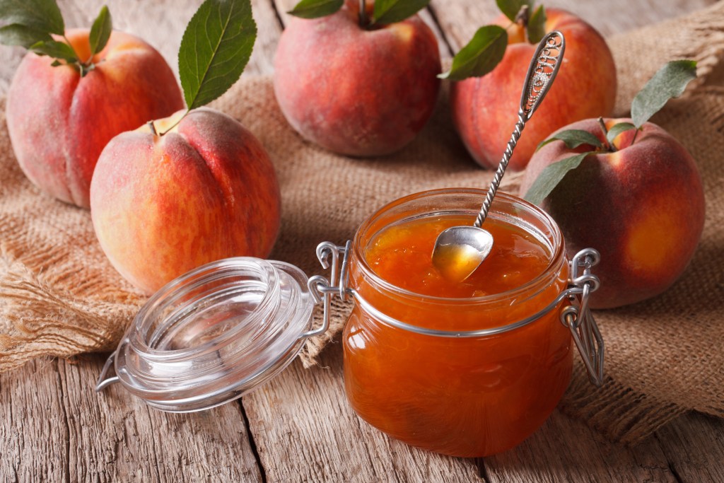 Jar of peach jam with spoon surrounded by fresh Chilton County peaches on rustic wooden surface