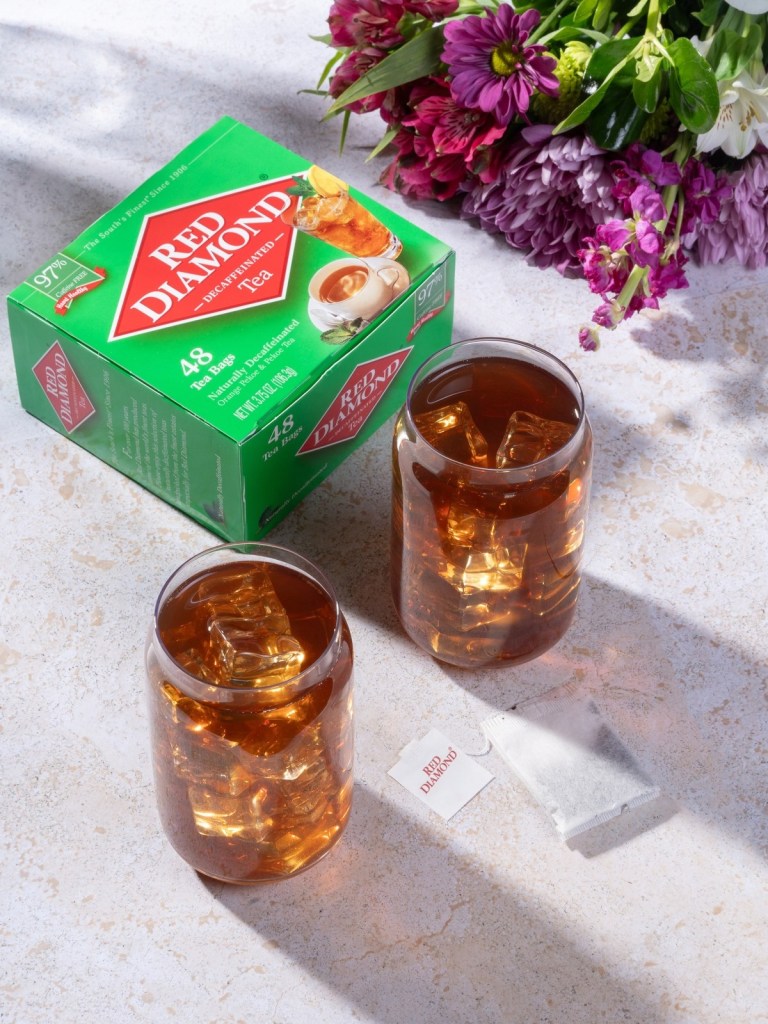 Box of Red Diamond Decaffeinated Tea with two glasses of iced tea, tea bag, and flowers on a light stone surface