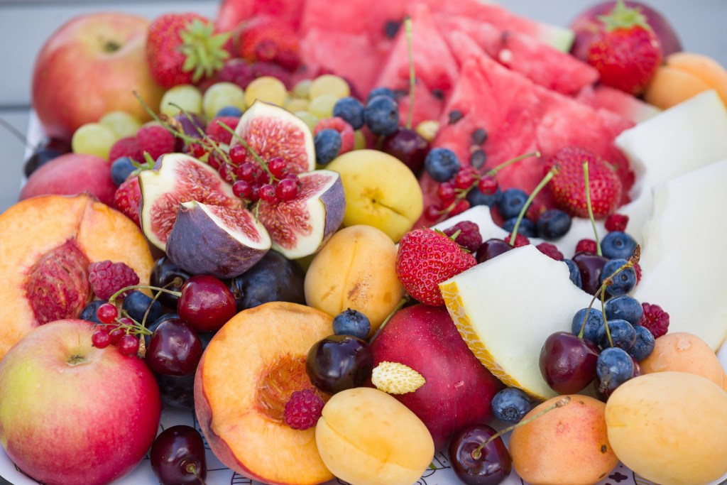 Assorted fresh summer fruits including Chilton County peaches, watermelon slices, figs, berries, cherries, and melon, displayed in a vibrant arrangement