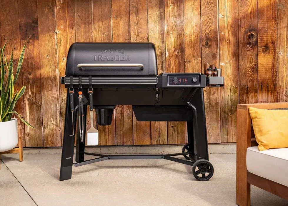 Traeger Woodridge WiFi Grill displayed on a patio with wooden wall, grill tools, and digital temperature display