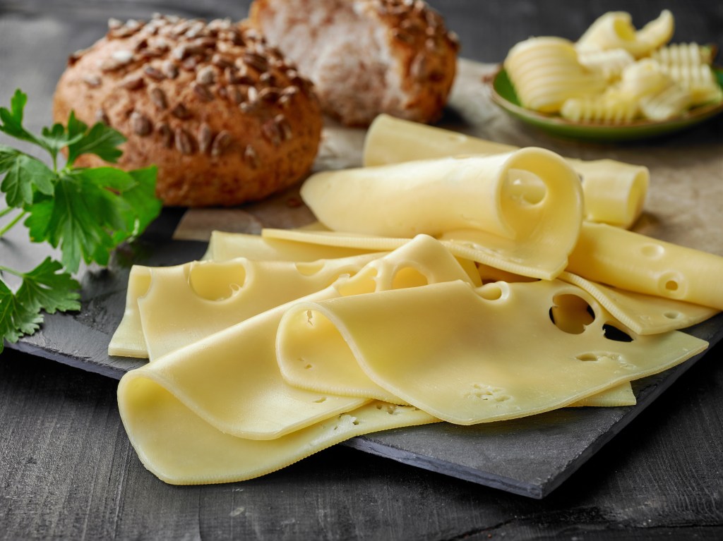 Thin slices of Swiss cheese on slate board with seeded bread rolls and butter curls