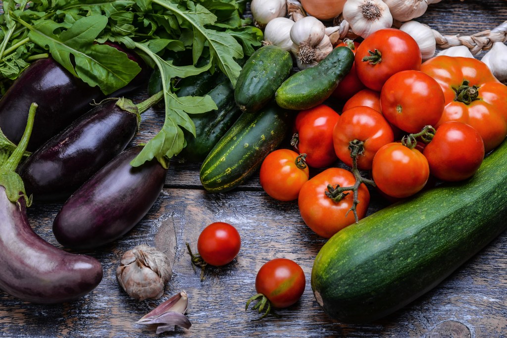 Assortment of fresh summer vegetables including tomatoes, cucumbers, eggplants, zucchini, garlic, and arugula on rustic wood
