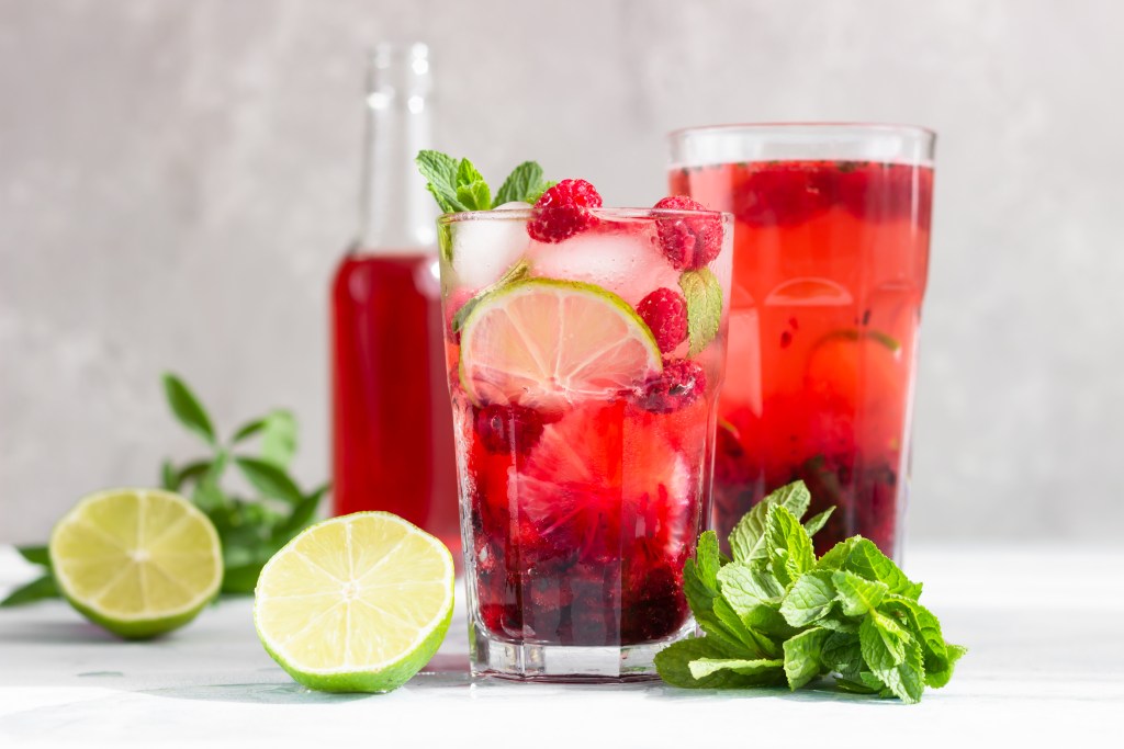 lass of red berry iced tea with lime wheel, fresh raspberries, and mint garnish, alongside lime halves and mint leaves, with a pitcher of berry tea in the background