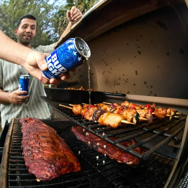 Person pouring Bud Light beer over grilled chicken kabobs while ribs cook on the grill at a backyard BBQ