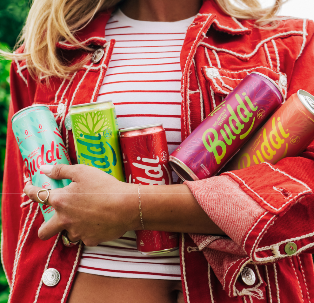 A woman wearing a red striped top and red jacket holds multiple vibrant cans of Buddi THC-infused seltzer in assorted flavors and colors.
