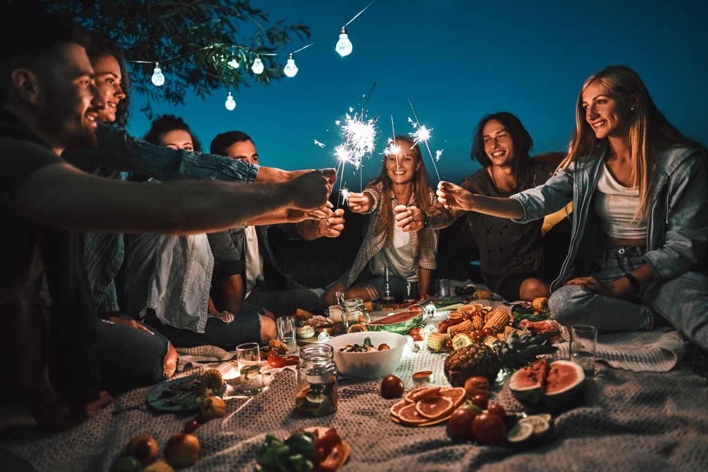 Group of young adults enjoying a festive Fourth of July picnic at dusk with sparklers, fresh fruits, vegetables, and summer foods under string lights