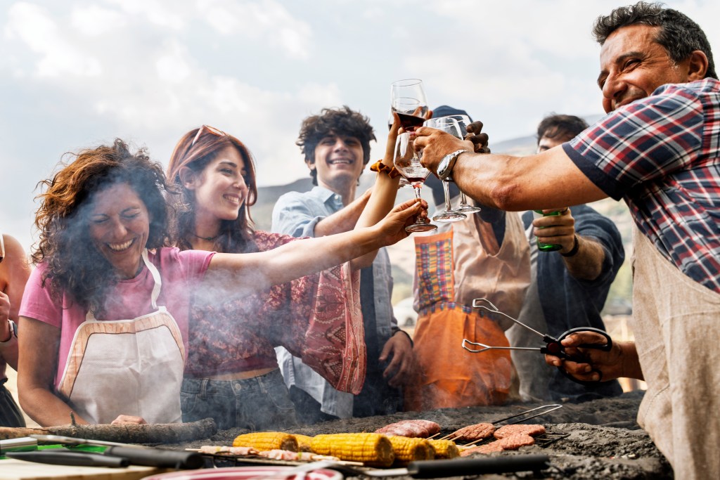 Group of friends toasting with wine and beer at an outdoor barbecue with corn and burgers on the grill