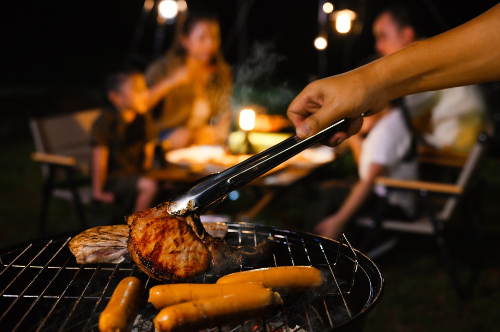 Hand grilling pork chop and sausages on a barbecue grill with blurred family in the background at night