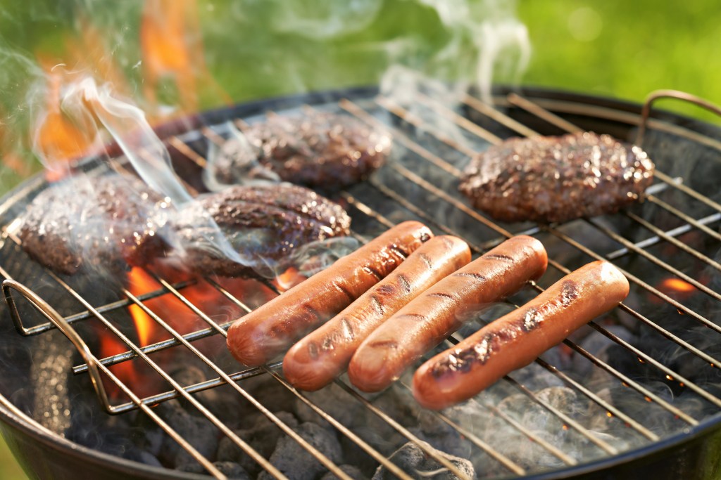 Hot dogs and hamburger patties sizzling on a charcoal grill with flames