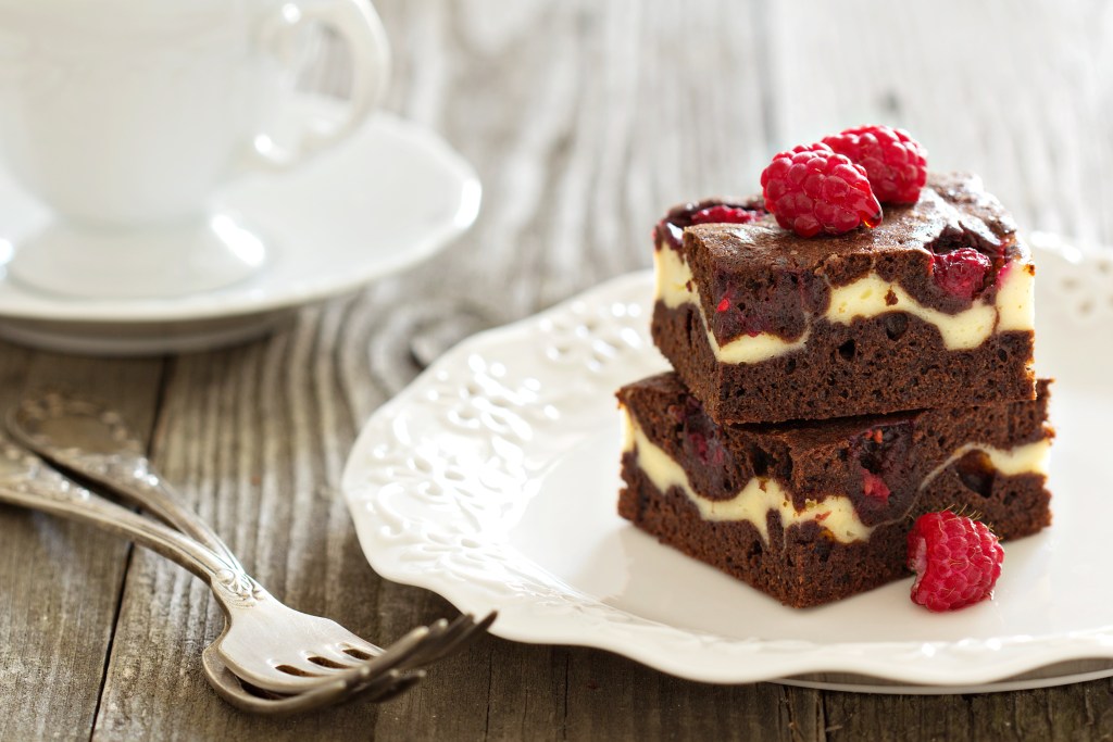 Two stacked chocolate brownies with cheesecake swirls and raspberries on a white decorative plate, served with vintage silverware on a rustic wooden table