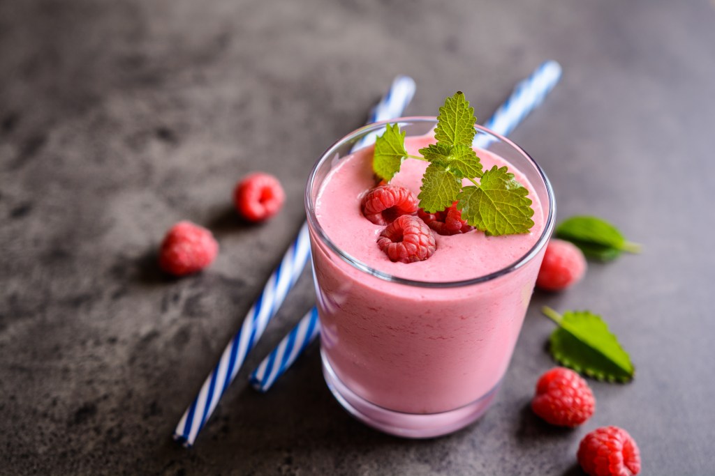 Glass of pink raspberry smoothie topped with fresh raspberries and mint leaves, surrounded by striped straws and scattered berries on a dark stone surface