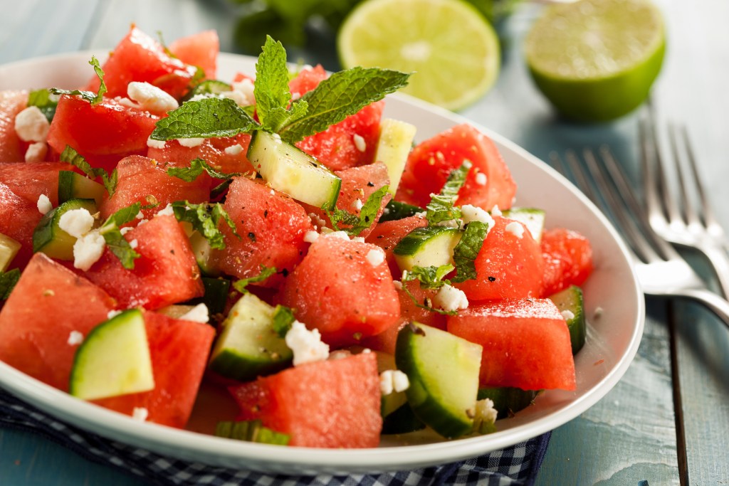 Close-up of a watermelon and cucumber salad garnished with crumbled feta cheese, fresh mint, and black pepper, served on a white plate