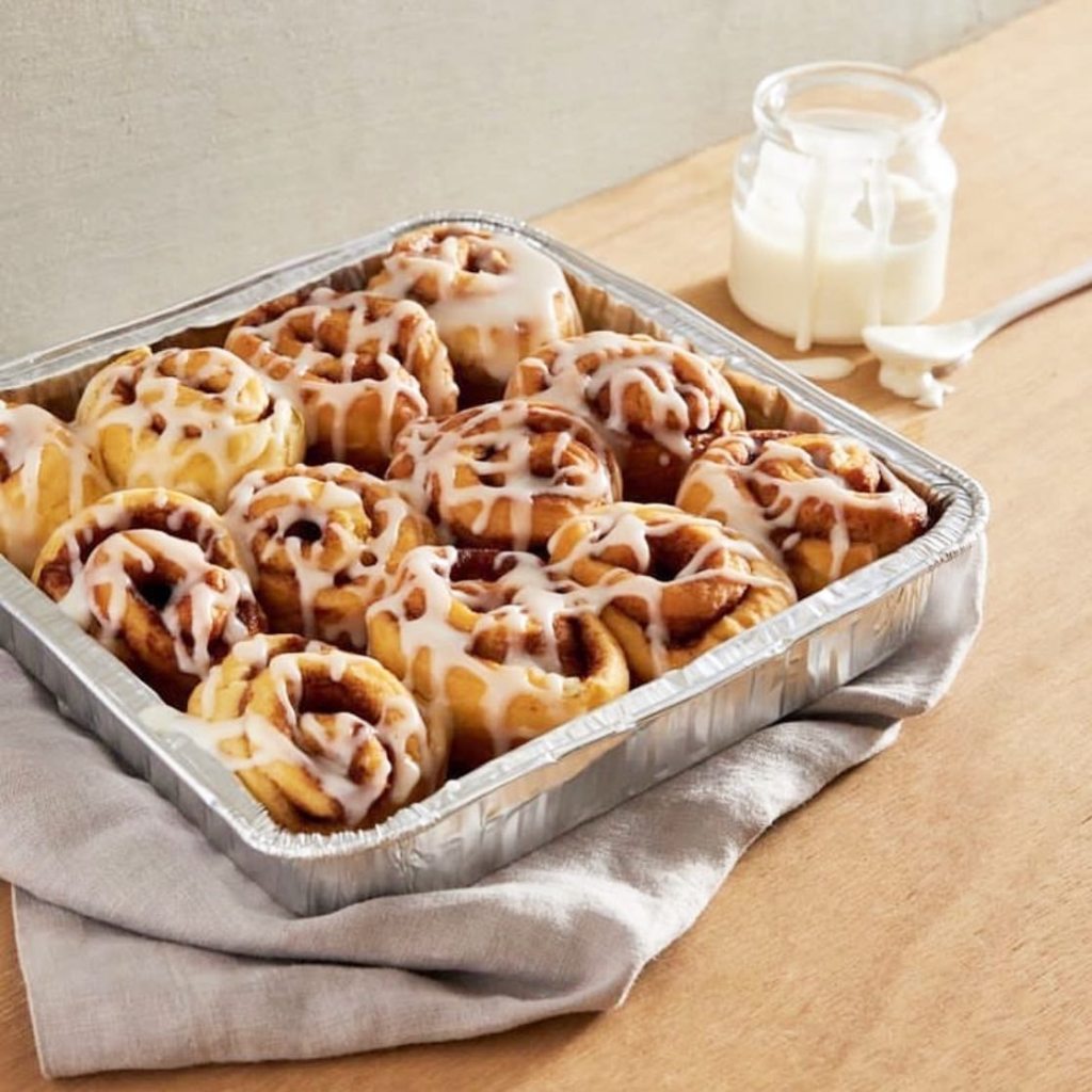 Tray of Millie Ray’s cinnamon rolls drizzled with white icing, placed on a wooden table with a jar of icing in the background