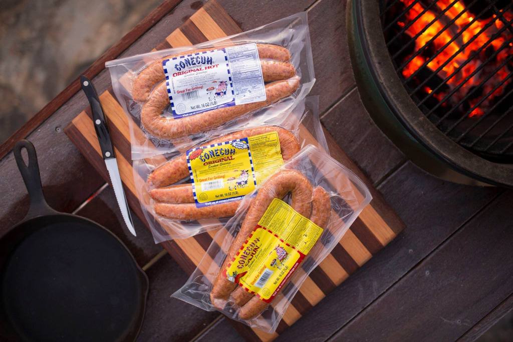 Three vacuum-sealed packs of Conecuh Sausage placed on a cutting board next to a grill and cast iron skillet