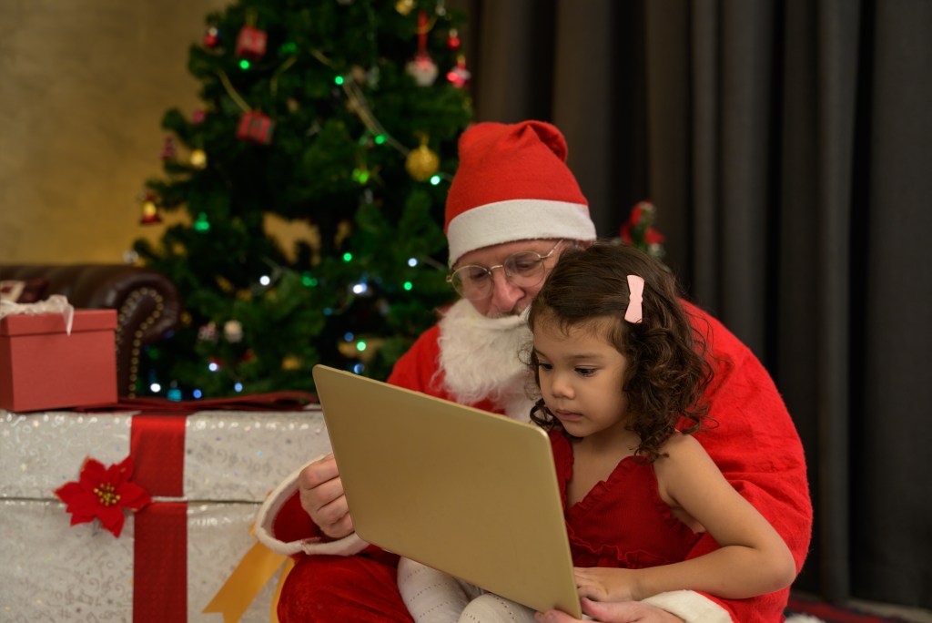 Santa Claus sitting with a young girl looking at a laptop in front of a decorated Christmas tree