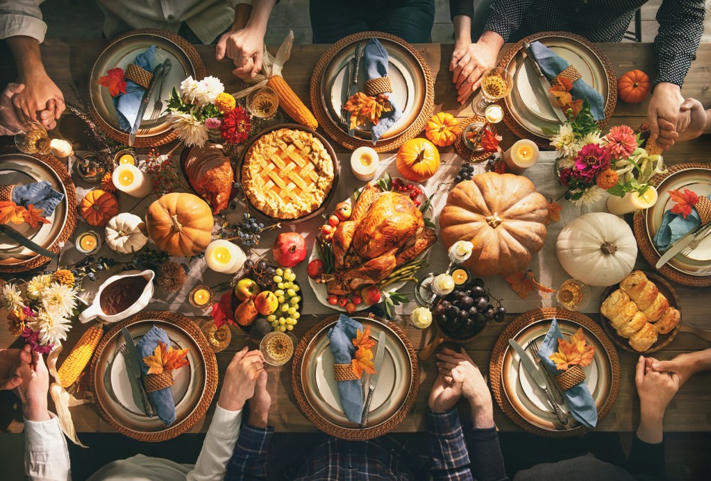 Overhead view of a Thanksgiving table with roast turkey, pie, pumpkins, candles, and a family holding hands in prayer
