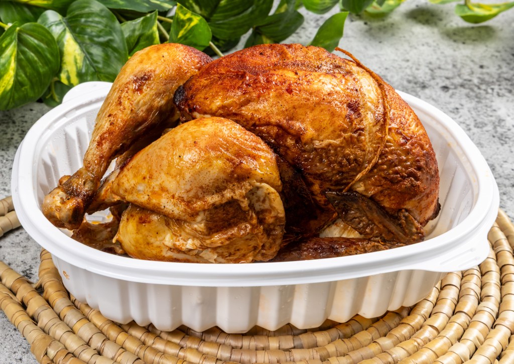 Golden-brown whole roasted chicken served in a white takeout container on a woven placemat
