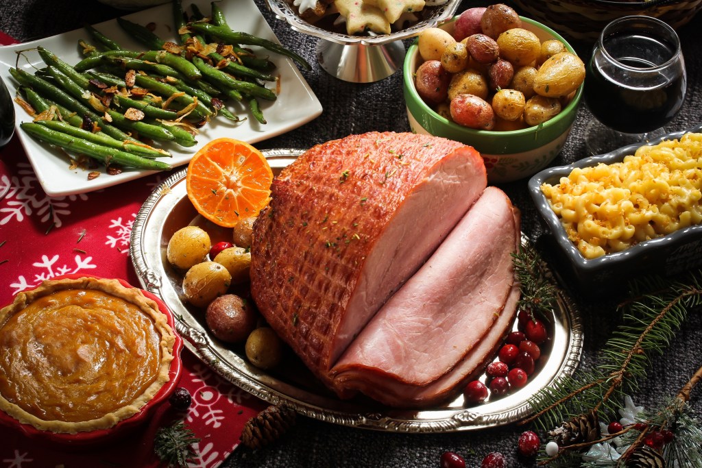 Sliced glazed ham on a holiday table with green beans, roasted potatoes, macaroni and cheese, and sweet potato pie