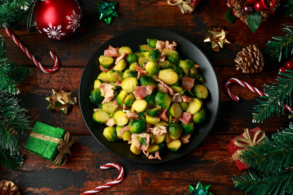 Bowl of cooked Brussels sprouts with bacon surrounded by Christmas decorations on a wooden table