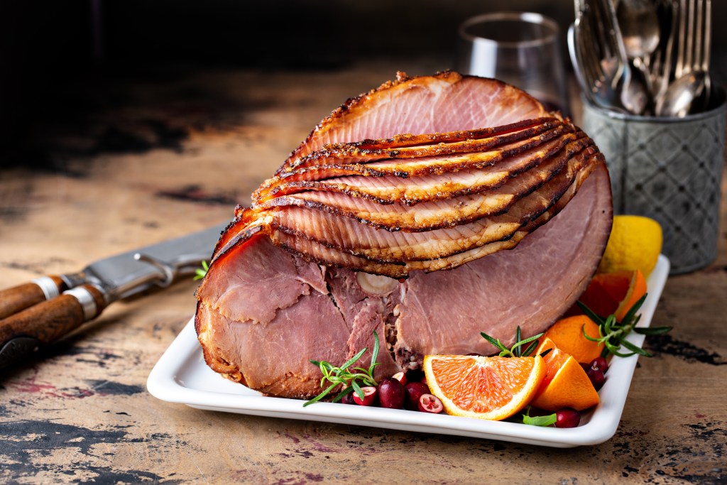 Spiral-sliced glazed ham with caramelized crust, garnished with orange slices, cranberries, and rosemary on a white platter