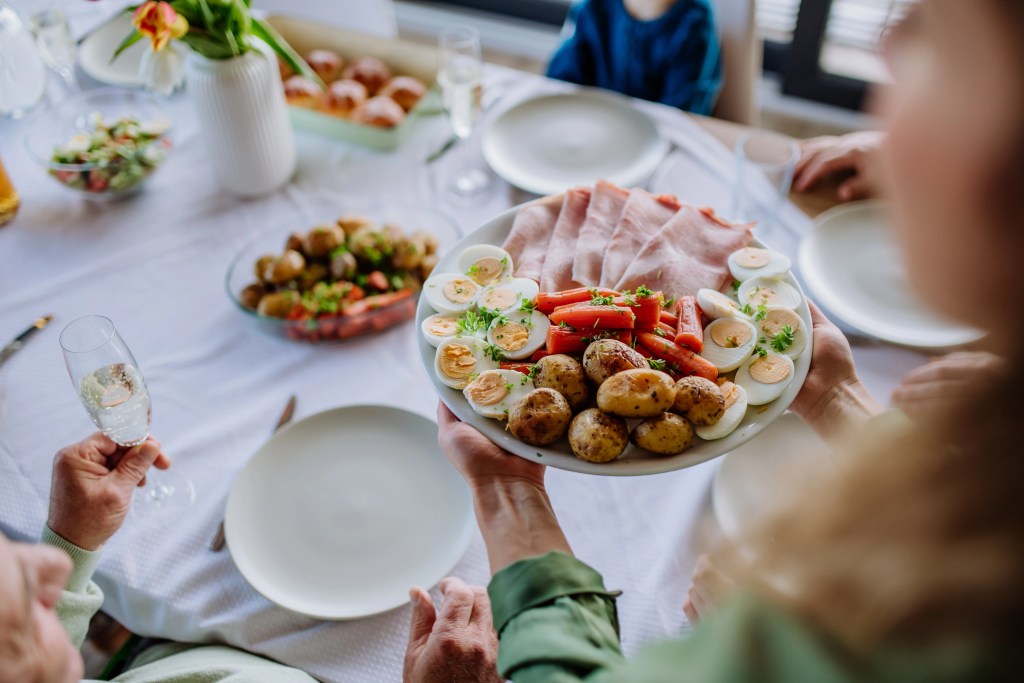 Close-up of a person serving a festive holiday platter with deviled eggs, ham slices, roasted potatoes, and vegetables at a decorated dining table