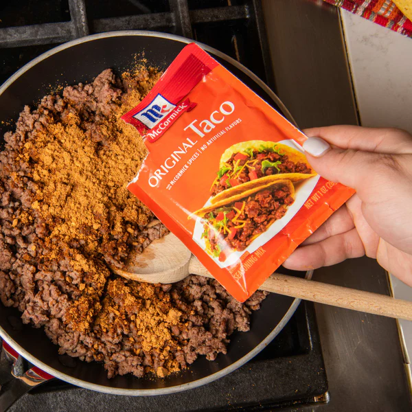 Hand pouring McCormick Original Taco seasoning packet into a skillet of browned ground beef on a stovetop