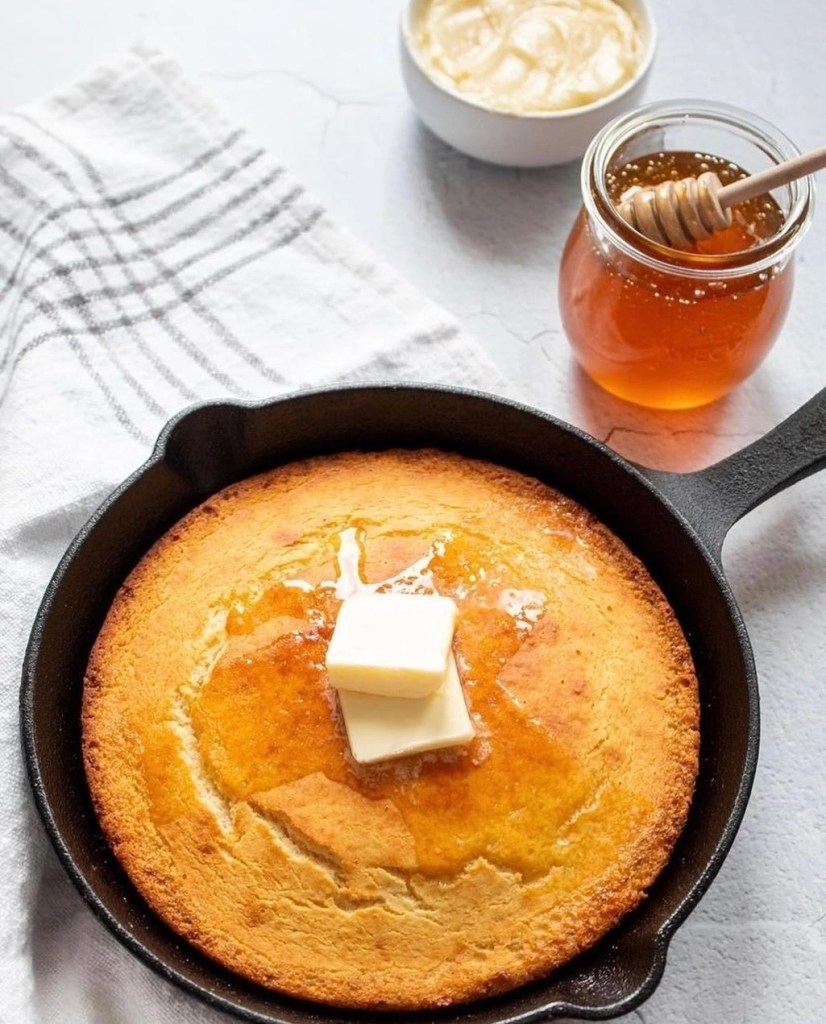 Golden Southern cornbread baked in a cast iron skillet topped with melting butter, served with a jar of honey on a light kitchen table