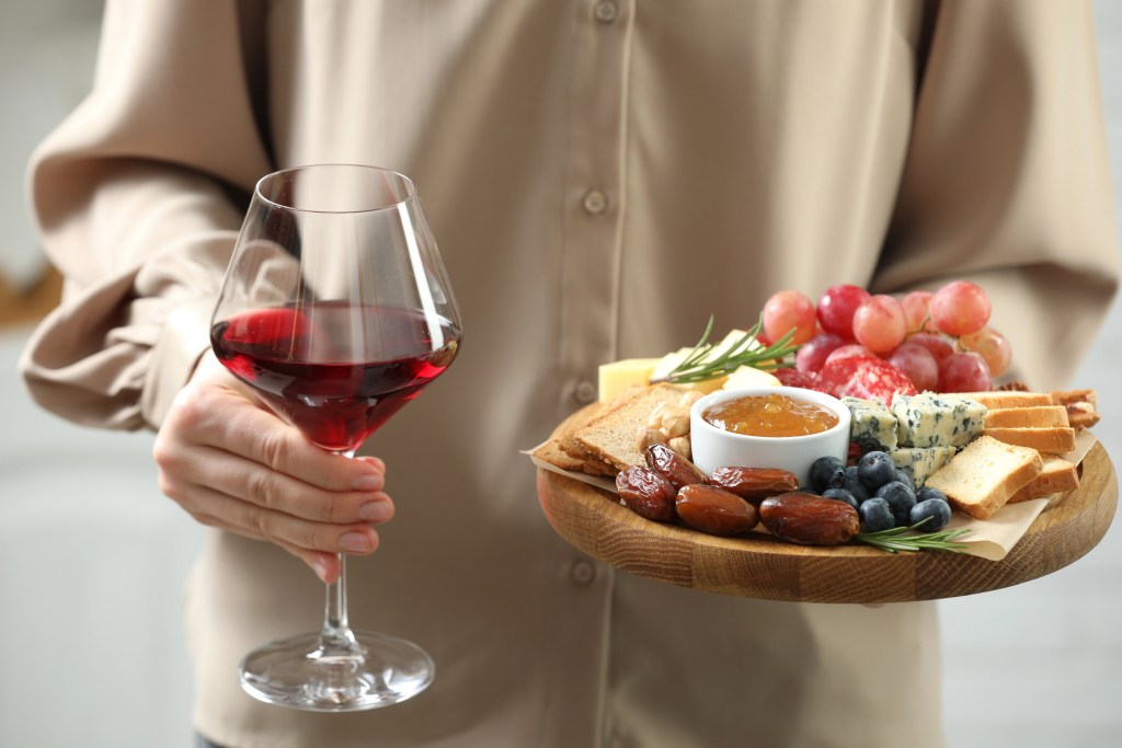 Person holding a glass of red wine and a wooden charcuterie board with brie, blue cheese, grapes, berries, crackers, dates, and honey