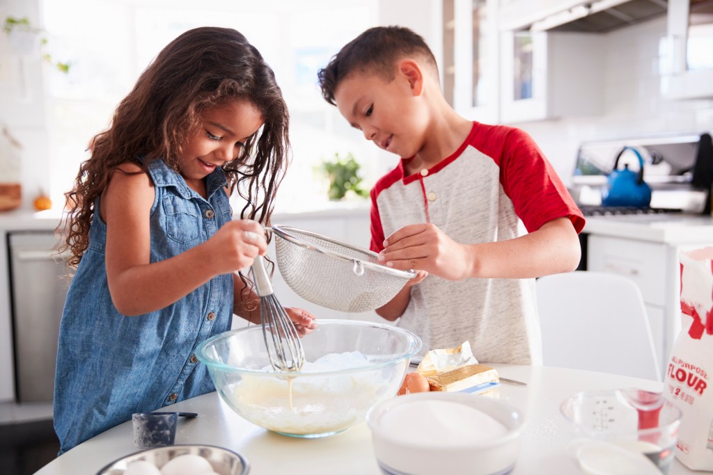 Two children baking together in a bright kitchen, mixing batter and sifting flour while preparing Valentine’s desserts at home