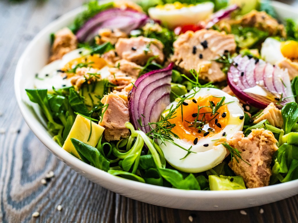 Classic tuna salad plate with chunks of tuna, boiled eggs, fresh greens, red onion, and avocado in a white bowl
