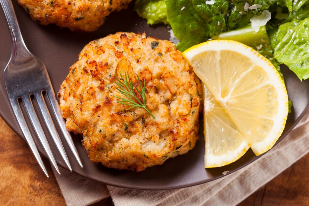 Close up of a golden brown crab cake garnished with dill and served with a lemon wedge and green salad on a brown plate