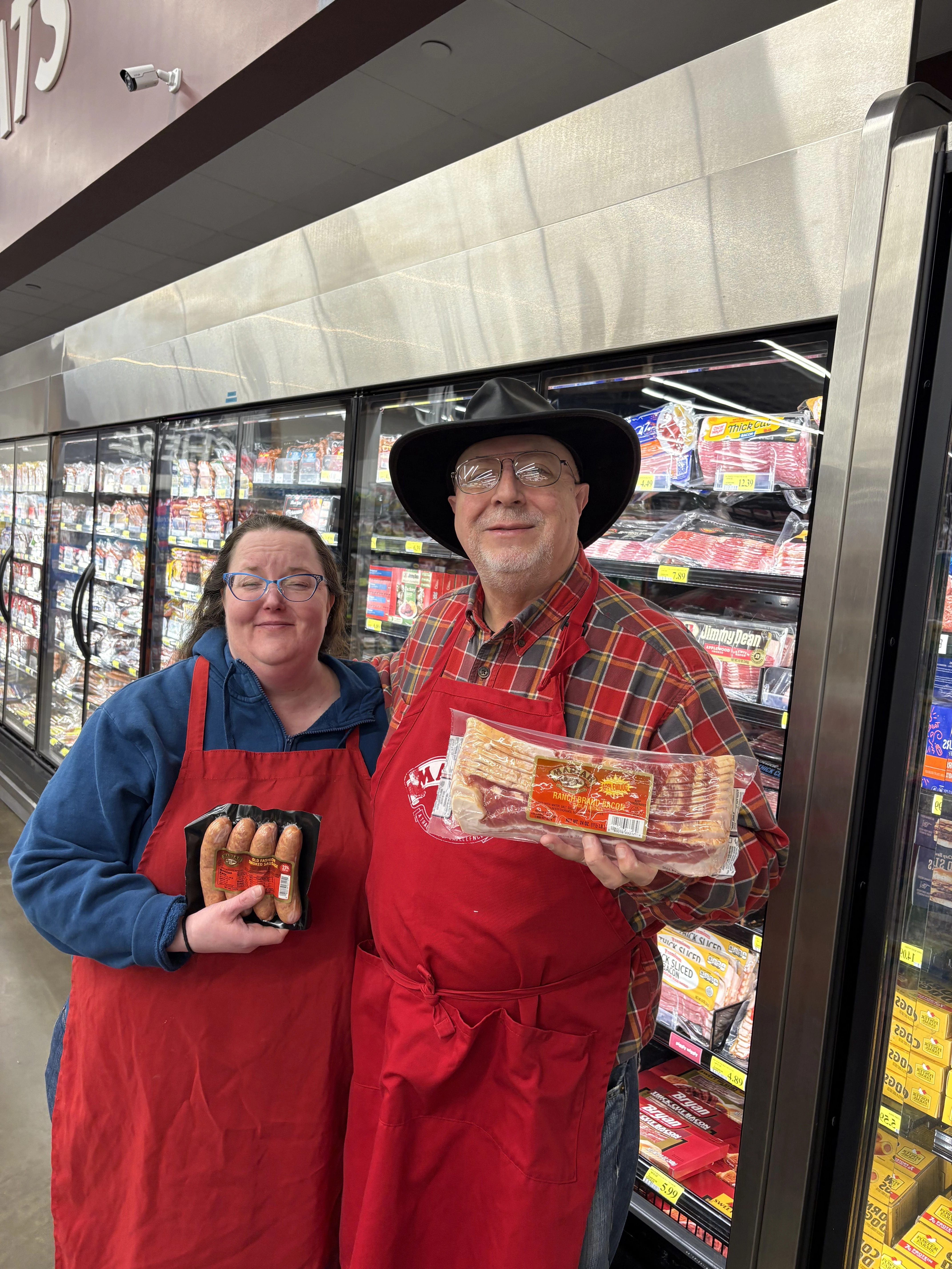 Two Piggly Wiggly employees in red aprons holding Mariah Premium bacon and smoked sausage during an in-store food tasting event in Birmingham