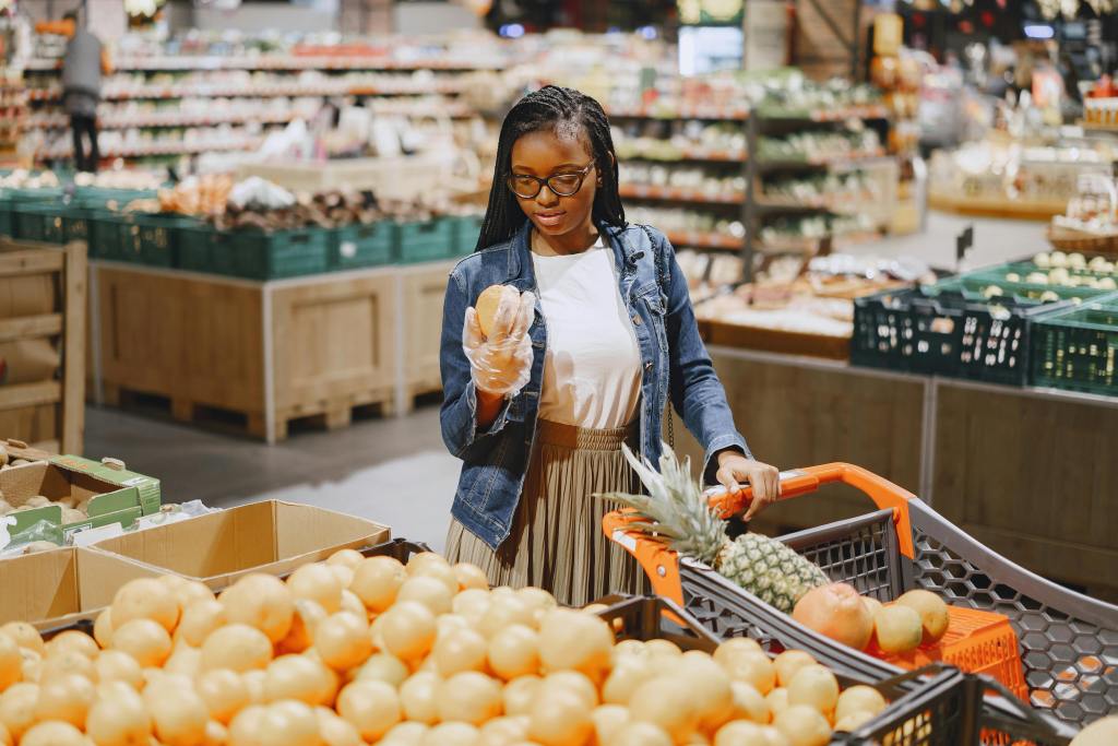 Woman wearing gloves selecting fresh fruit in a grocery store produce section with a shopping cart