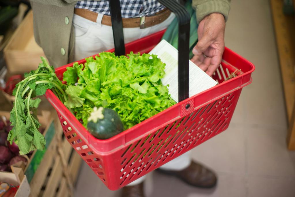 Shopper holding a red grocery basket filled with fresh leafy greens and vegetables inside a neighborhood grocery store